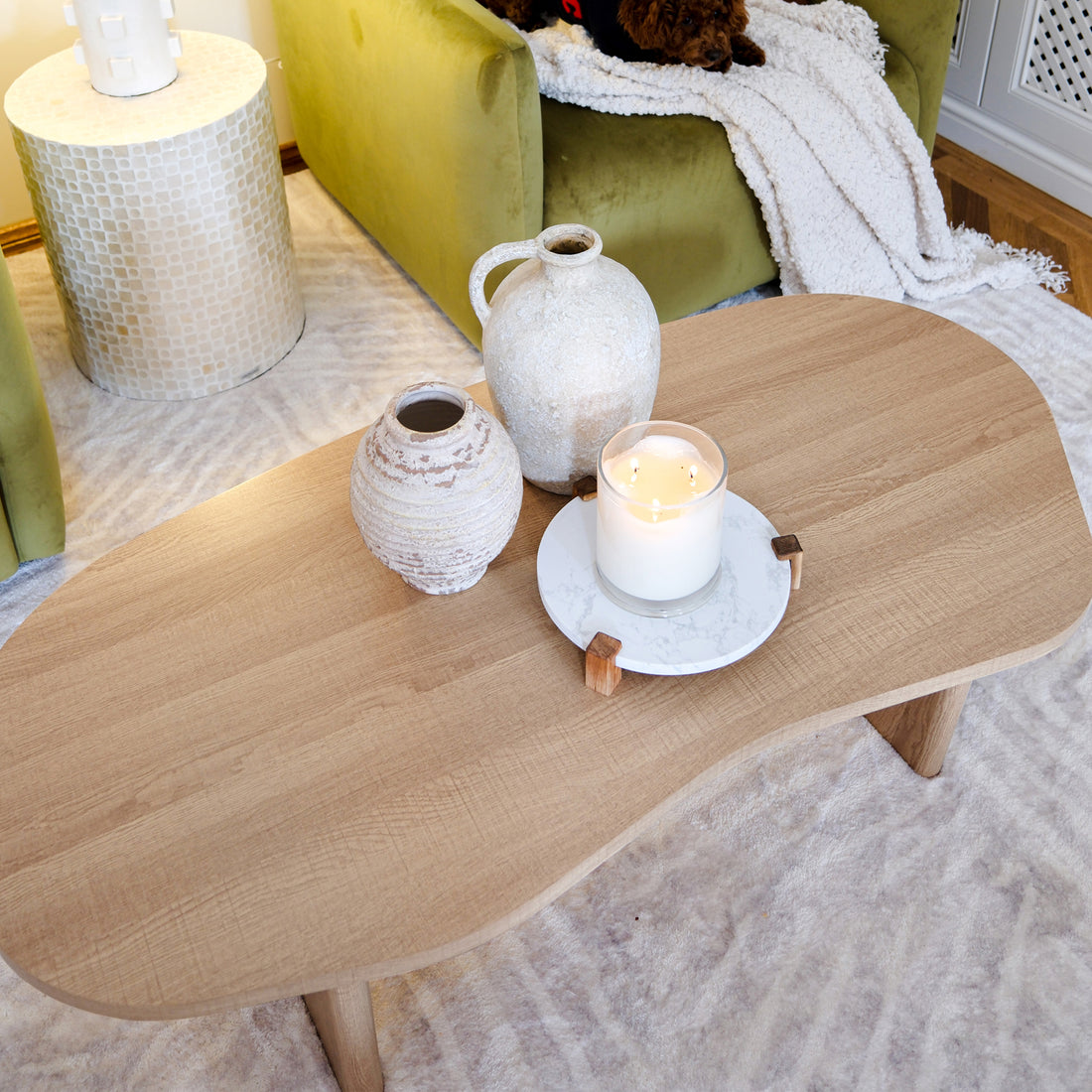 Nicole 120cm Oval Coffee Table in Natural with soft wood finish, featuring two ceramic vessels, a three-wick candle on a tray, set against a cozy interior backdrop with a plush chair and side table.