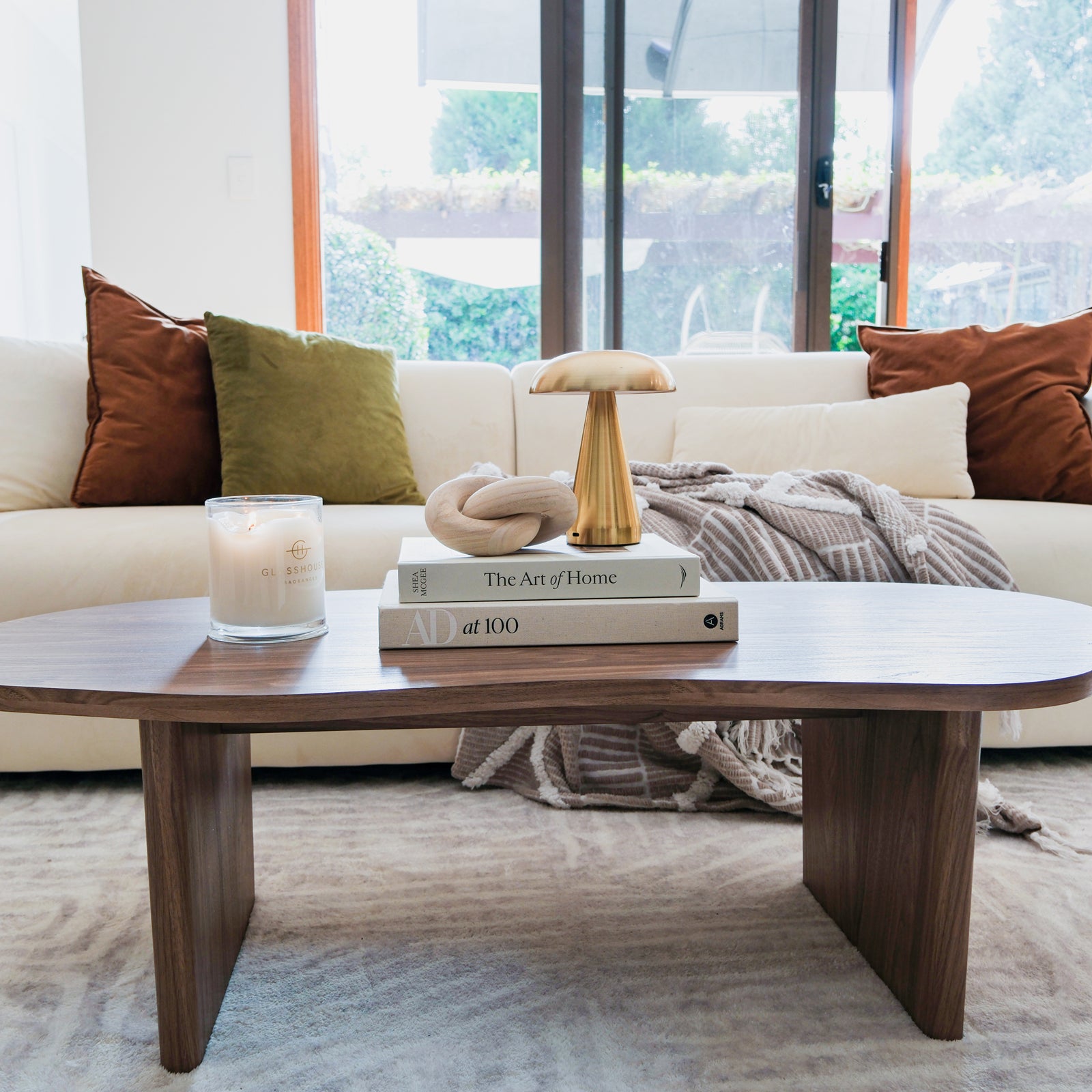 Nicole 120cm Coffee Table with Organic Curve in Walnut, featuring a rich walnut finish, holds a lit candle, coffee-table books, a wooden knot, and a brass mushroom lamp, against a cozy living-room backdrop.