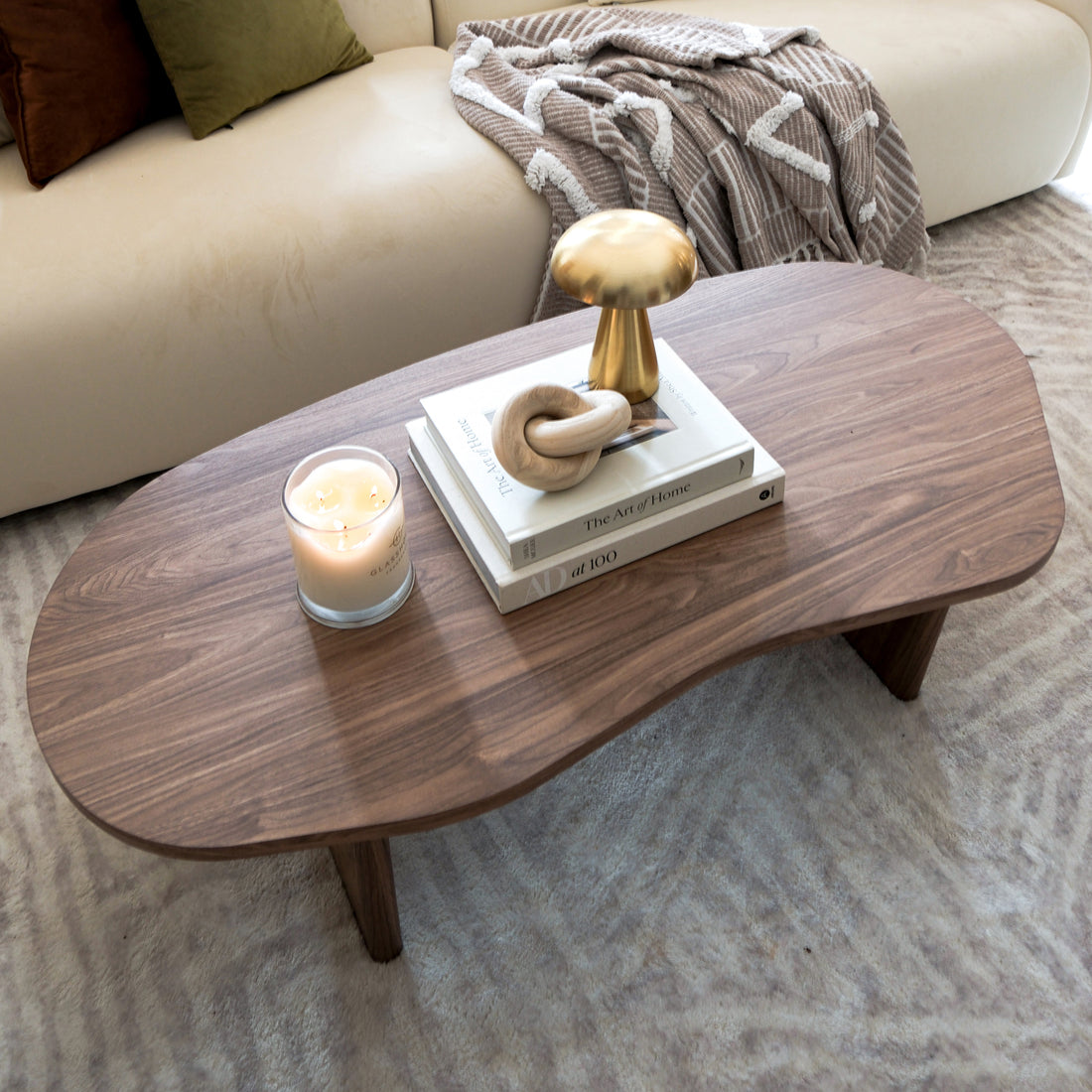 Nicole 120cm Coffee Table with Organic Curve in Walnut, featuring a smooth walnut finish, holds a lit candle, books, a wooden sculpture, and a gold decorative object, exuding modern elegance.