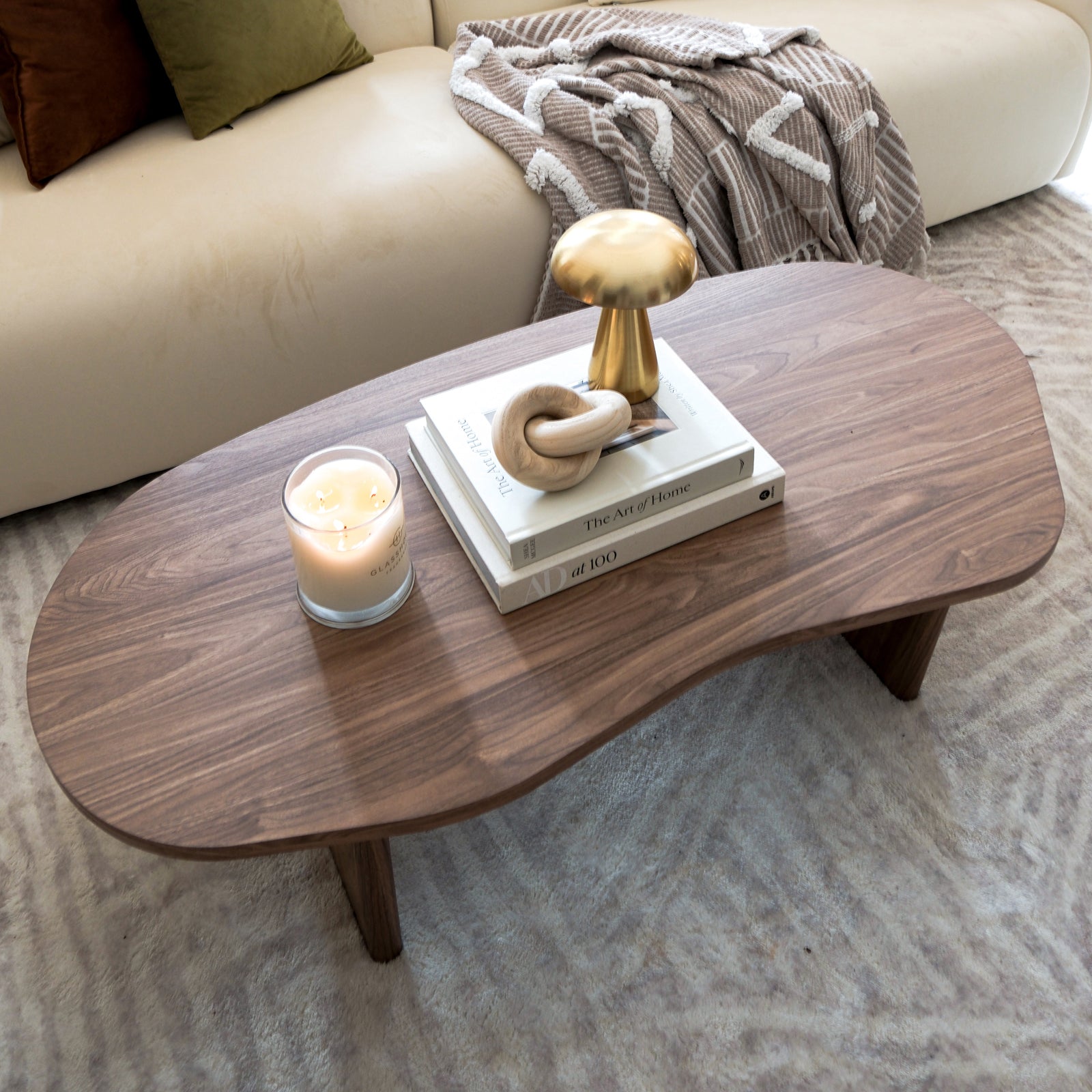Nicole 120cm Coffee Table with Organic Curve in Walnut, featuring a smooth walnut finish, holds a lit candle, books, a wooden sculpture, and a gold decorative object, exuding modern elegance.