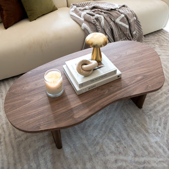 Nicole 120cm Coffee Table with Organic Curve in Walnut, featuring a smooth walnut finish, holds a lit candle, books, a wooden sculpture, and a gold decorative object, exuding modern elegance.