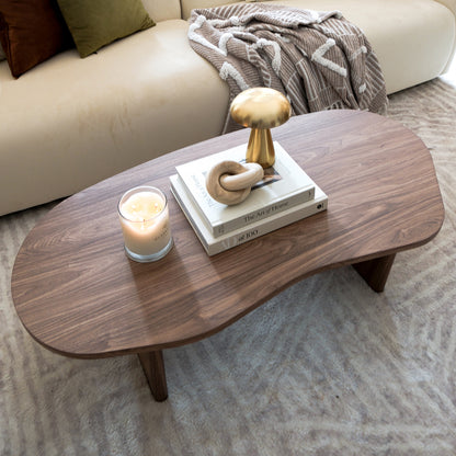 Nicole 120cm Coffee Table with Organic Curve in Walnut, featuring a smooth walnut finish, holds a lit candle, books, a wooden sculpture, and a gold decorative object, exuding modern elegance.