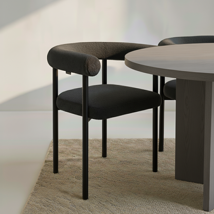 Ohio Curved Boucle Dining Chair Set of 2 in Black featuring a padded seat and curved backrest with black metal legs, tucked beside a round wooden table on a woven area rug.