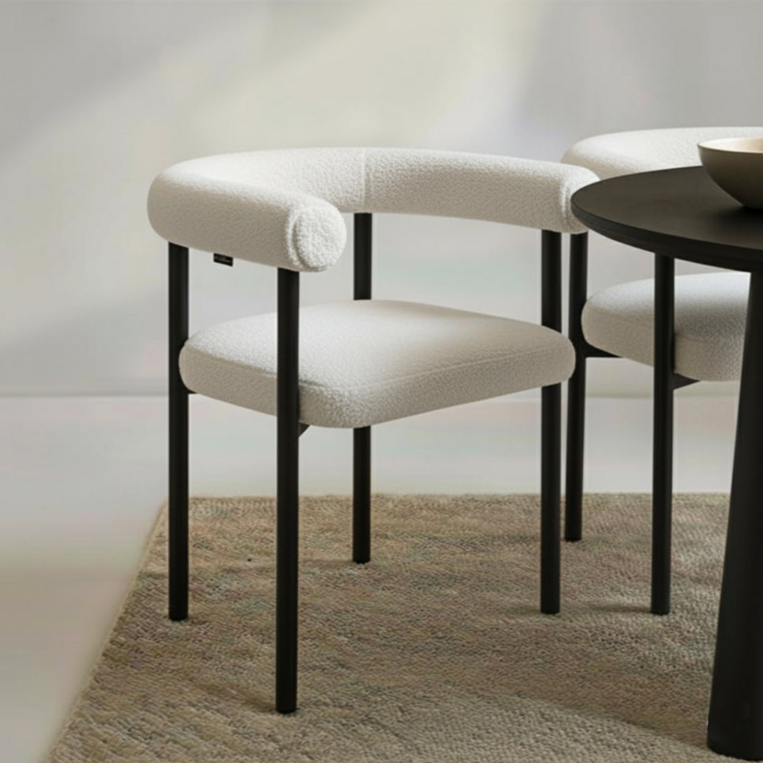 Ohio Curved Bouclé Dining Chair Set of 2 in Cream, featuring a rounded backrest and cushioned seat in off-white bouclé, with sleek black metal legs, arranged on a natural-fiber rug.