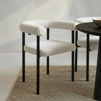 Ohio Curved Bouclé Dining Chair Set of 2 in Cream, featuring a rounded backrest and cushioned seat in off-white bouclé, with sleek black metal legs, arranged on a natural-fiber rug.