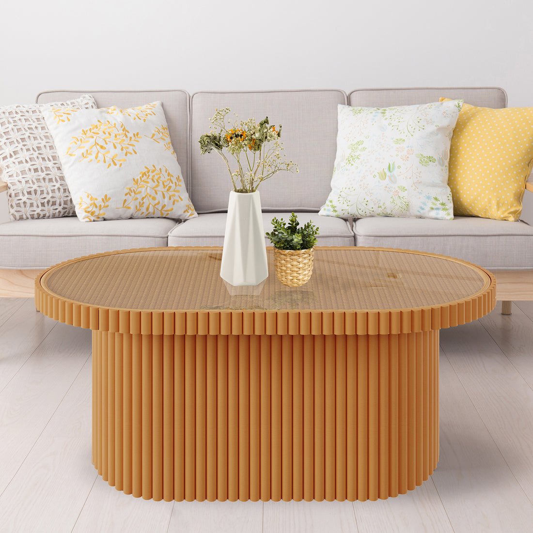 Olive Natural Rattan Side Coffee Table - Honey, featuring an oval design with a tempered glass top, displayed in a cozy setting with a plant, white pillow with gold leaves, and a yellow pillow.