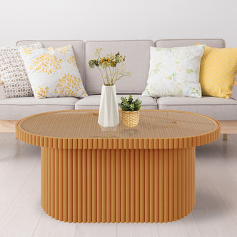 Olive Natural Rattan Side Coffee Table - Honey, featuring an oval design with a tempered glass top, displayed in a cozy setting with a plant, white pillow with gold leaves, and a yellow pillow.