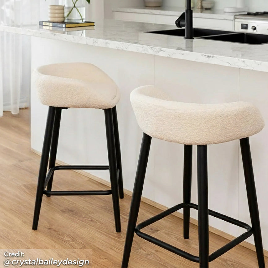 Peyton Bouclé Fabric Bar Stool Set of 2 in Cream features curved cream bouclé seats on black metal legs with footrest, elegantly tucked under a white marble-style countertop, enhancing modern kitchen aesthetics.