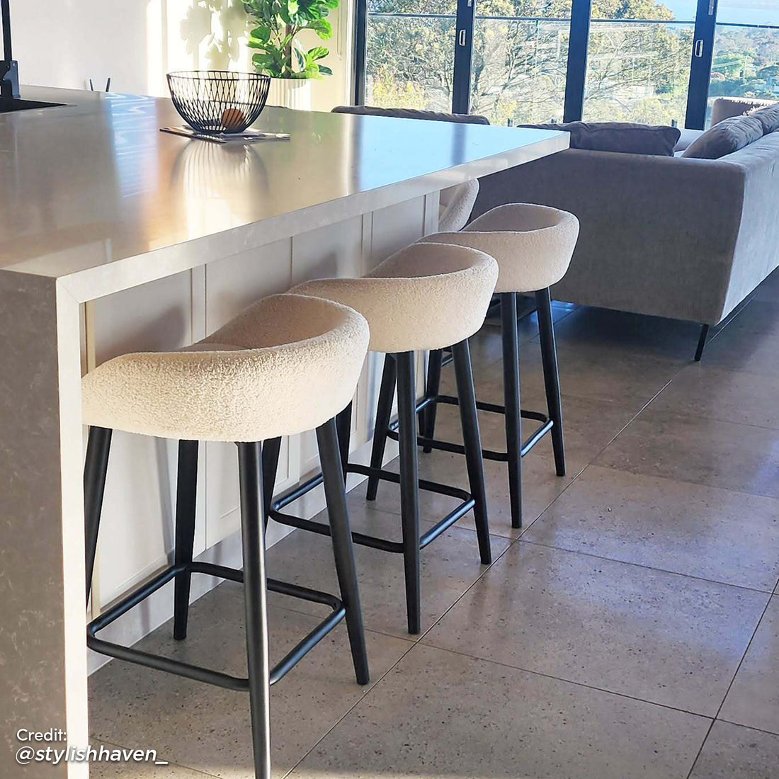 Peyton Bouclé Bar Stool Set of 2 in Cream, featuring soft upholstery and slender black legs with footrests, complements the modern kitchen's waterfall-edge island and open-plan design.
