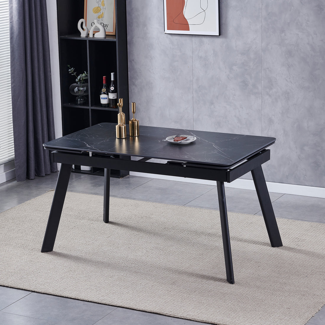 Rheed 197cm Extendable Ceramic Dining Table in Black with splayed legs on a woven rug, adorned with gold candleholders and food, set against a shelving unit and abstract art backdrop.