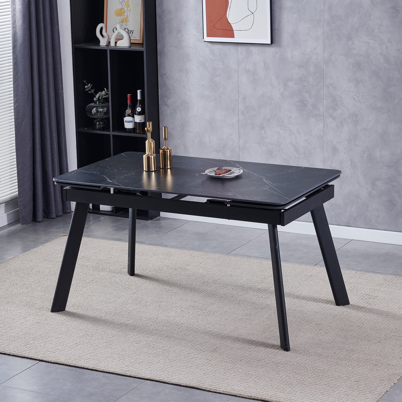 Rheed 197cm Extendable Ceramic Dining Table in Black with splayed legs on a woven rug, adorned with gold candleholders and food, set against a shelving unit and abstract art backdrop.