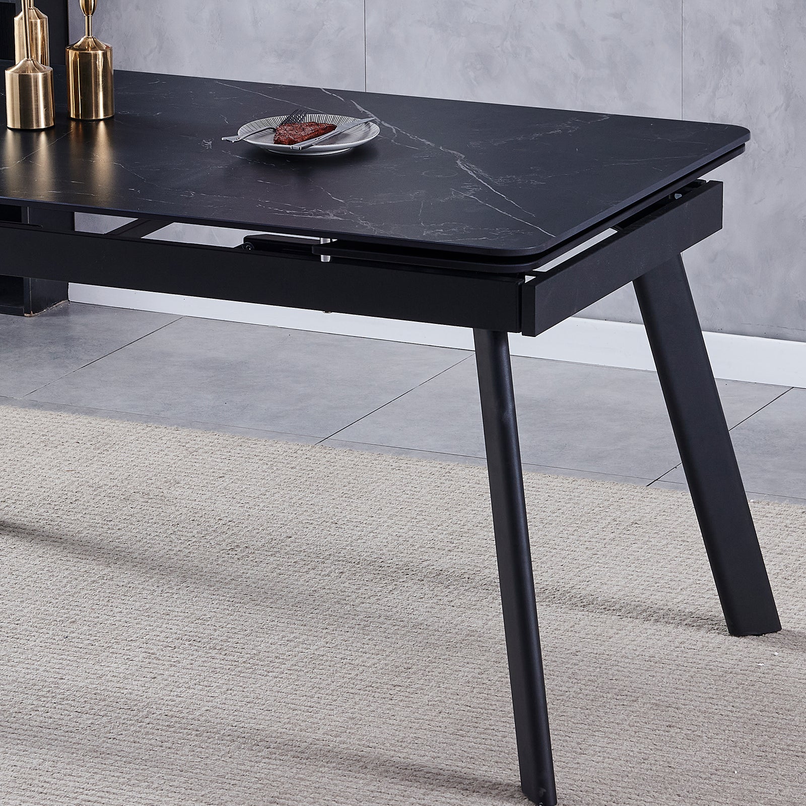 Rheed 197cm Extendable Ceramic Dining Table in Black with a faux-marble finish, splayed metal legs, and adorned with brass decorative vessels. A slice of meat and fork rest on a plate.
