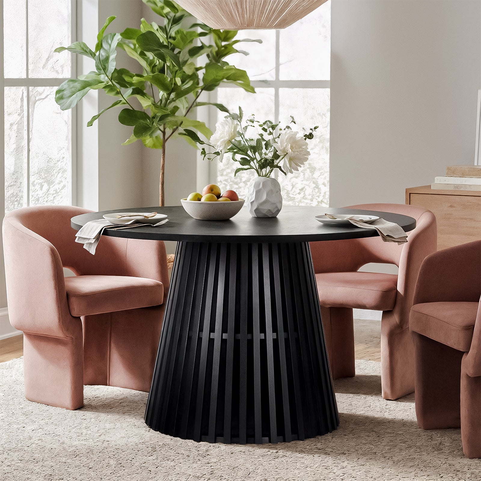 Riri 120cm Round Fluted Wood Dining Table in Black with a sculpted pedestal base, surrounded by blush-pink chairs, topped with fruit bowl, vase, and place settings, in a sunlit dining nook.