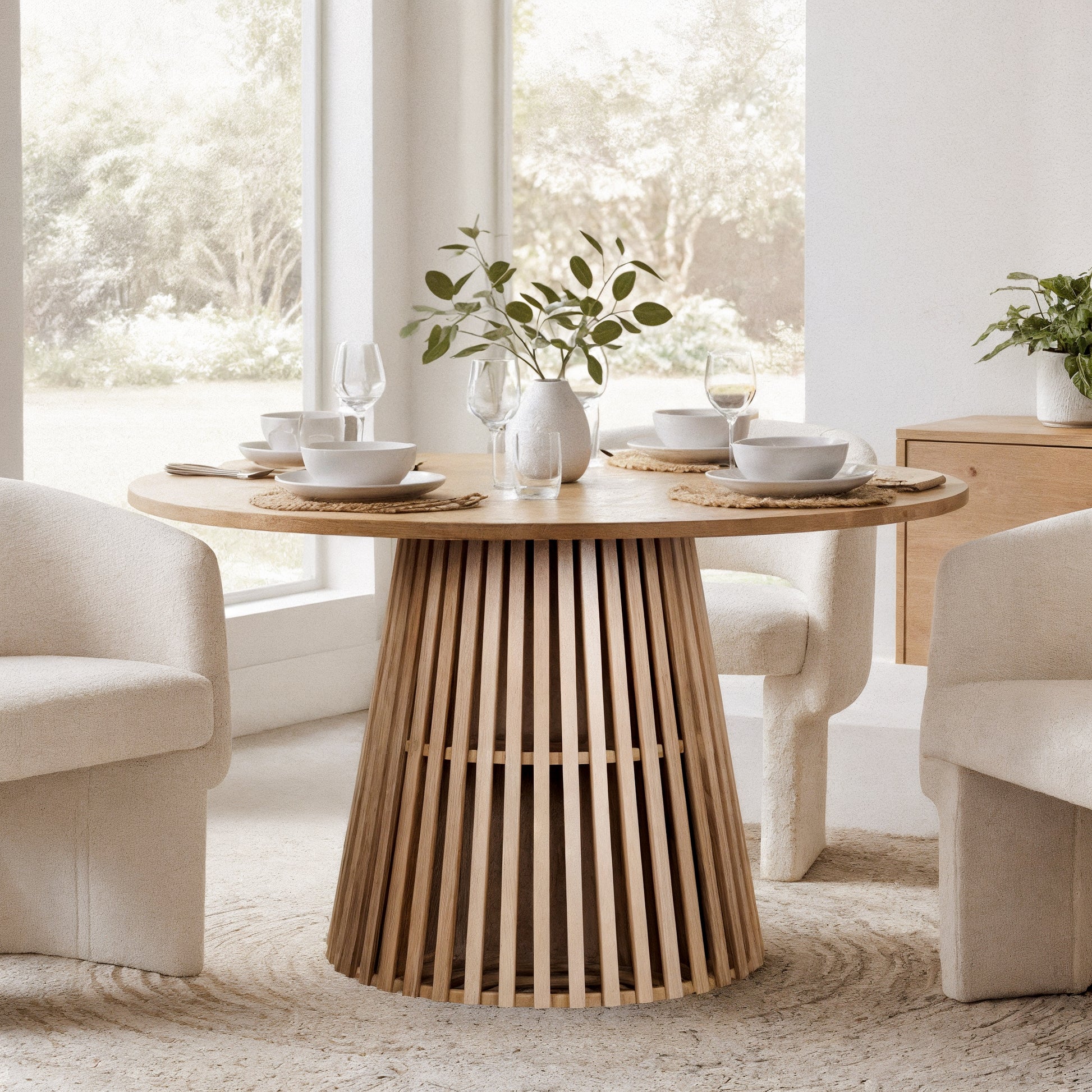 Riri 120cm Round Fluted Wood Dining Table in Oak, featuring a round top on a slatted pedestal base, set for four with white tableware, woven placemats, and a leafy ceramic vase centerpiece.