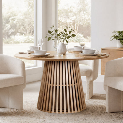 Riri 120cm Round Fluted Wood Dining Table in Oak, featuring a round top on a slatted pedestal base, set for four with white tableware, woven placemats, and a leafy ceramic vase centerpiece.