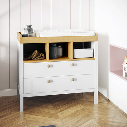 Rosella Solid Pine Baby Change Table with Drawers in White, featuring two drawers, three cubbies with folded cloths, and a changing mat on top, against a white-paneled wall on hardwood flooring.