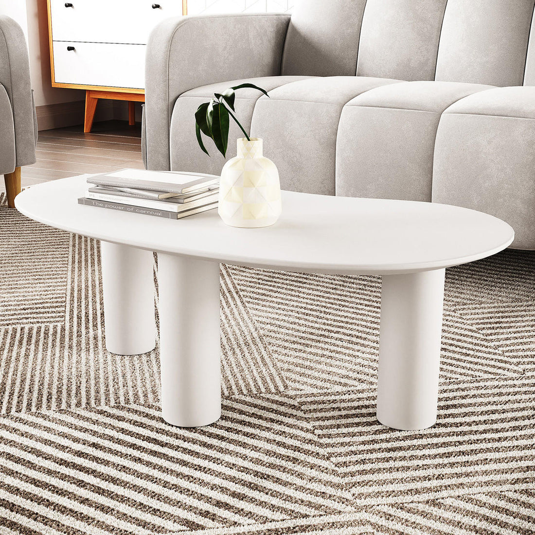 Sallie Cashew Coffee Table with Pillar Legs in White, featuring an oval shape, chunky cylindrical legs, with a ceramic vase and books on top, set on a textured, striped rug.