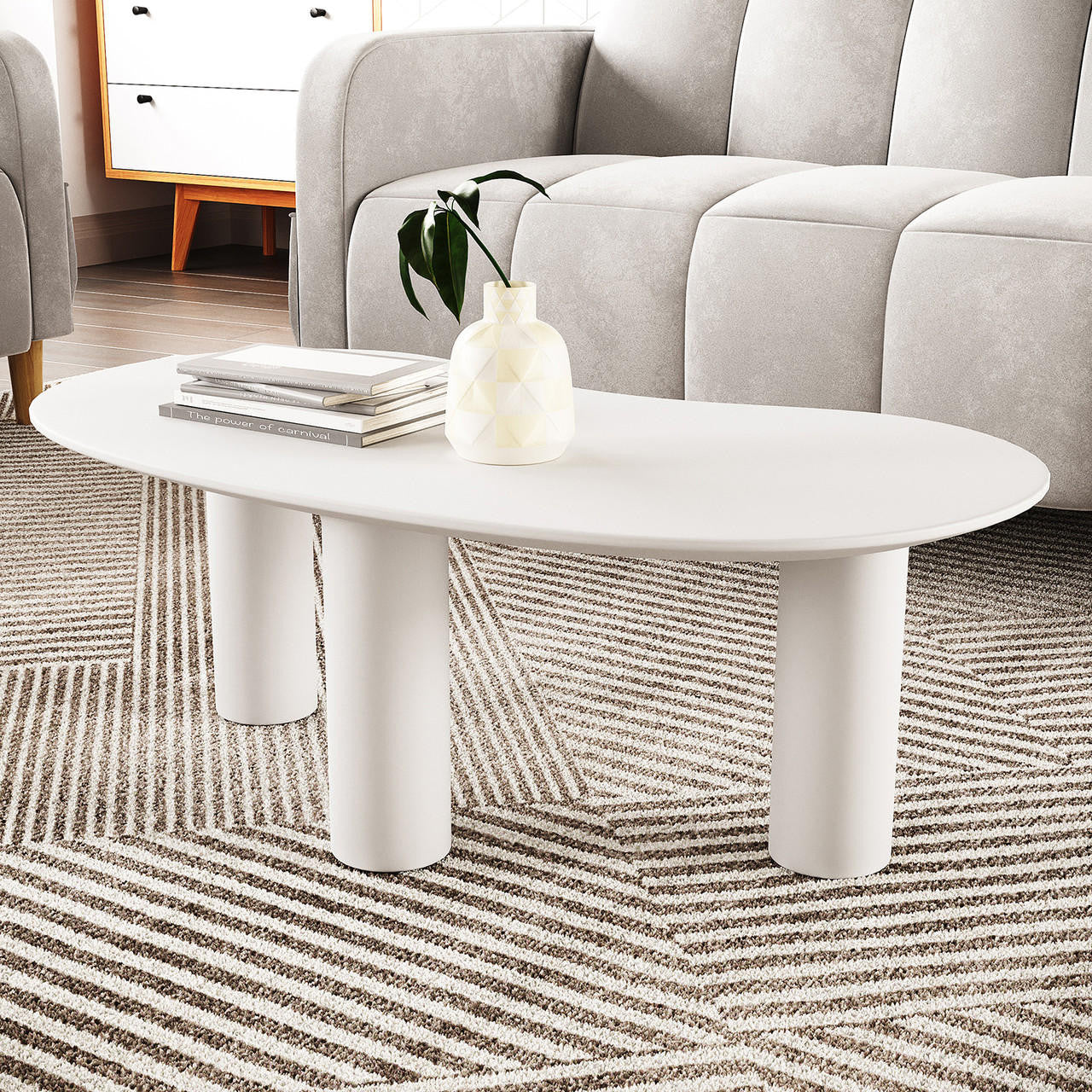 Sallie Cashew Coffee Table with Pillar Legs in White, featuring an oval shape, chunky cylindrical legs, with a ceramic vase and books on top, set on a textured, striped rug.