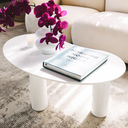 Sallie Cashew Coffee Table with Pillar Legs in White, featuring a sleek oval design, three cylindrical legs, and topped with a book, vase, and candle, set on a textured rug.