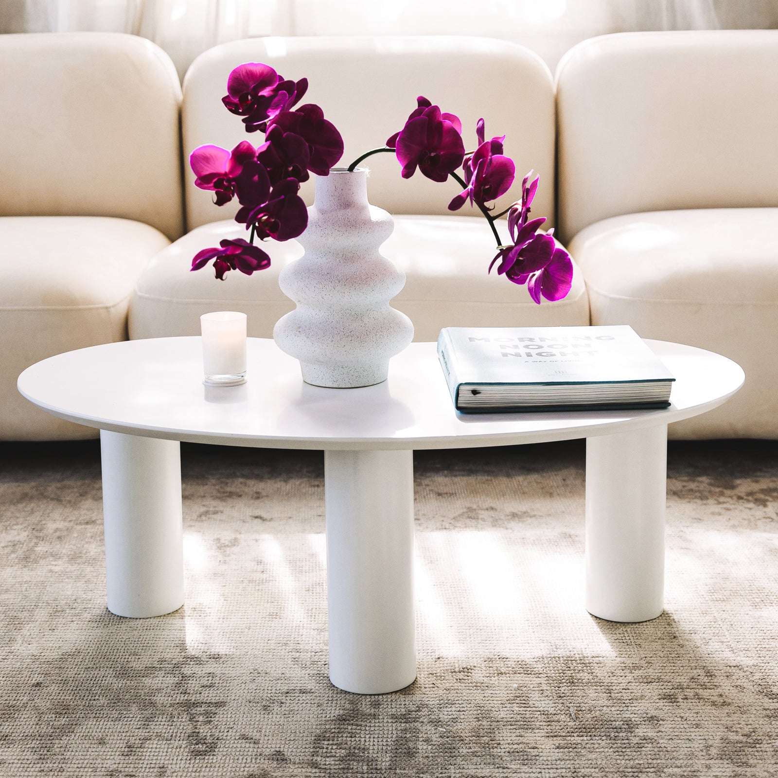 Sallie Cashew Coffee Table with Pillar Legs in White, on a rug, features a sculptural vase with magenta orchids, a votive candle, and a hardcover book, creating a modern, elegant setting.