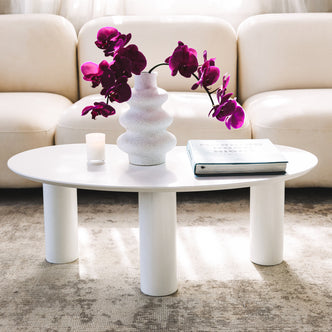 Sallie Cashew Coffee Table with Pillar Legs in White, on a rug, features a sculptural vase with magenta orchids, a votive candle, and a hardcover book, creating a modern, elegant setting.