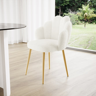 The Sally Scalloped Boucle Dining Chair Set of 2 features cream bouclé upholstery with a scalloped backrest and integrated arms, supported by sleek gold metal legs on a light wood floor.