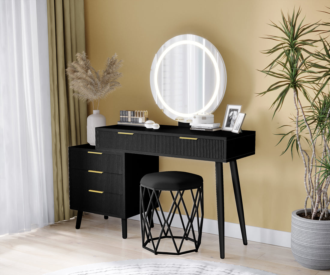 Serenity Makeup Vanity Table with LED Touch Mirror in Black, featuring ribbed drawers, gold handles, round backlit mirror, pampas grass vase, lipsticks, compacts, and a cushioned stool.