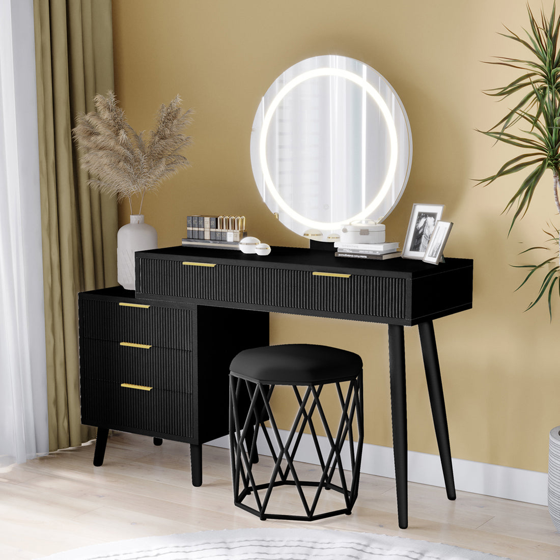 Serenity Makeup Vanity Table with LED Touch Mirror in Black, featuring ribbed drawers, gold handles, round backlit mirror, pampas grass vase, lipsticks, compacts, and a cushioned stool.