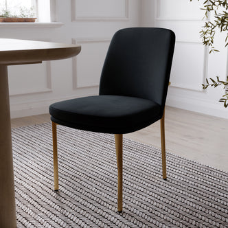 Shannon Velvet Dining Chair Set of 2 in Black features elegant black upholstery with slim gold-toned legs, showcased in a bright room with a striped rug, round table edge, and nearby plant.