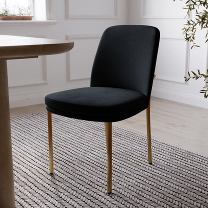 Shannon Velvet Dining Chair Set of 2 in Black features elegant black upholstery with slim gold-toned legs, showcased in a bright room with a striped rug, round table edge, and nearby plant.