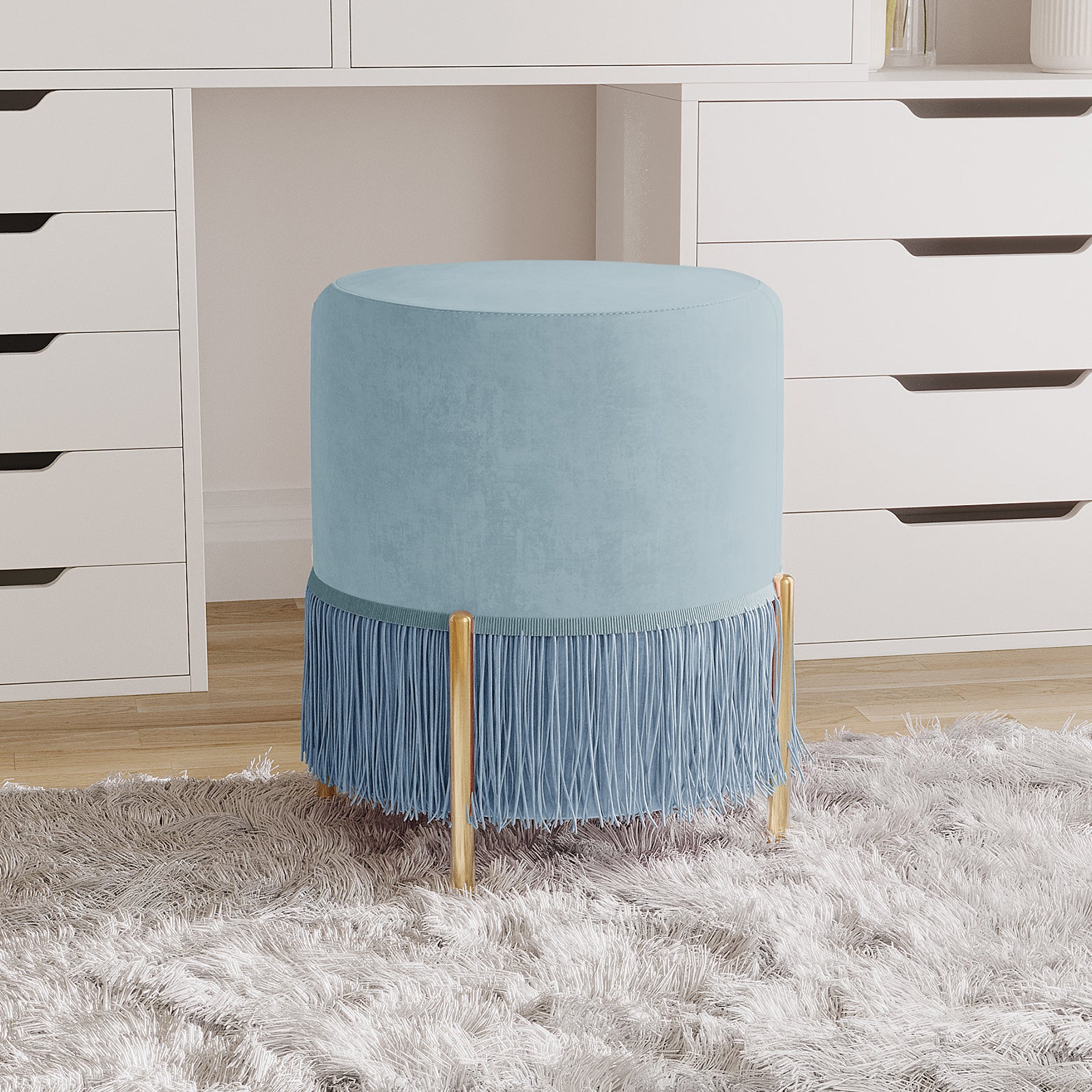 Stacey Tassled Velvet Ottoman Foot Stool in Blue, featuring plush blue upholstery, fringe tassels, and gold-tone legs, stands on an off-white shag rug in front of a white cabinet.