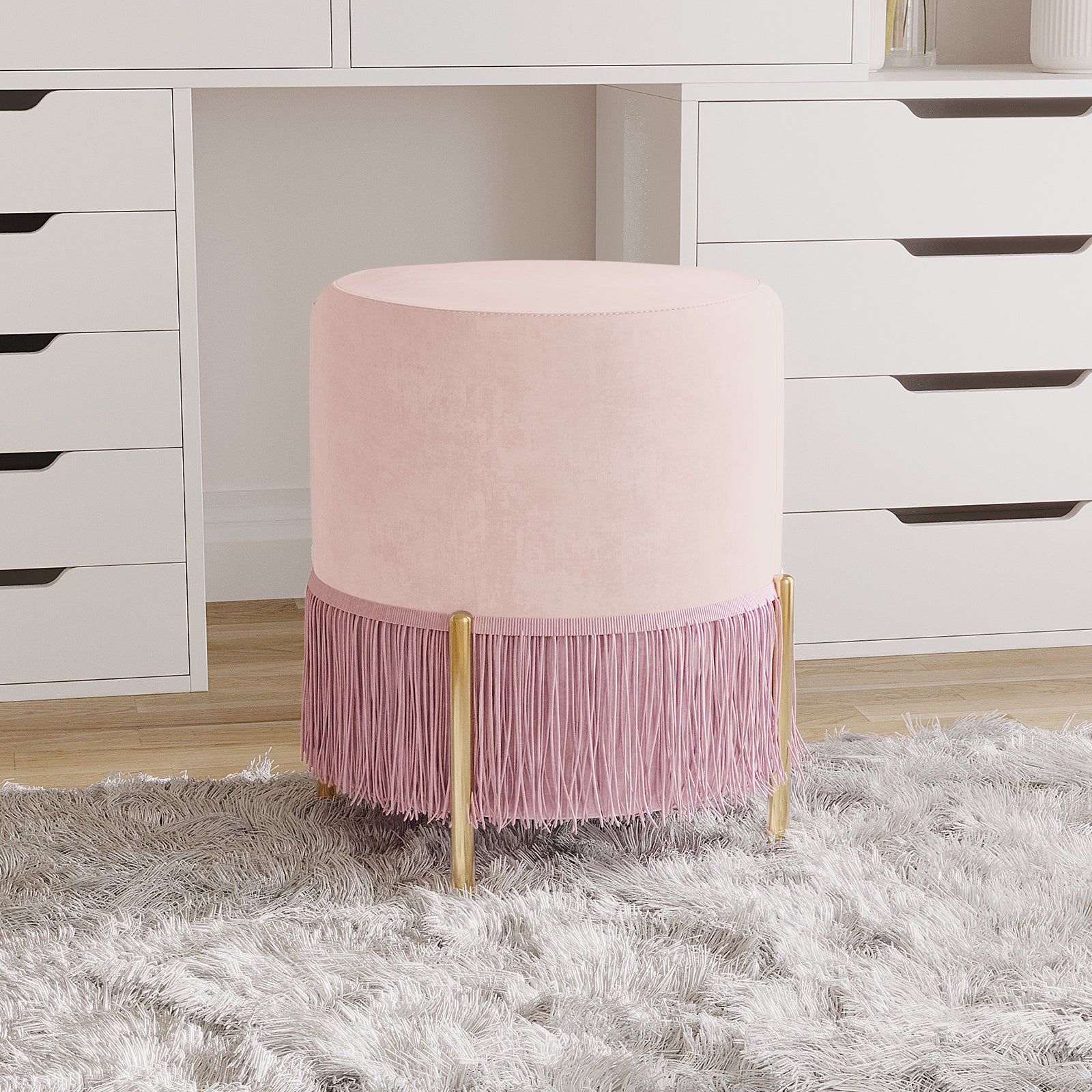 Stacey Velvet Foot Stool with Tassles in Light Pink, featuring blush-pink velvet upholstery, decorative fringe, and slender gold legs, sits on a shag rug near a white dresser, enhancing elegance and functionality.