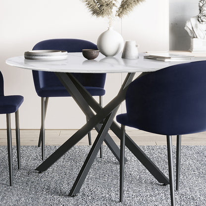Stoten Round Dining Table White Marble with a round marble top and black criss-cross legs, accompanied by navy chairs, a vase with dried grasses, and decor on a gray rug.
