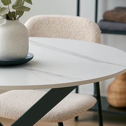 Stoten Round Dining Table White Marble features a marble-look top with dark metal legs, accompanied by a ceramic vase with green leaves, set in a minimalist modern interior.