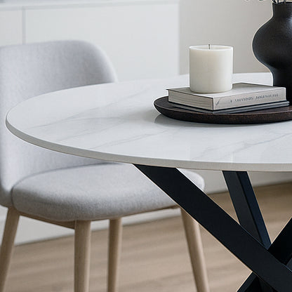 Stoten Round Dining Table White Marble with a marble-look top and black crisscross metal legs, paired with a wooden tray, books, candle, and black vase, set in a minimalist interior.