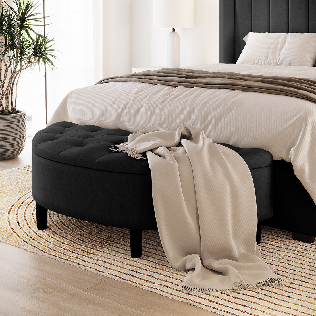 Amina Half Round Linen Fabric Ottoman in Black, tufted design, on a striped rug with a beige throw. Positioned in a modern bedroom corner, enhancing elegance and functionality.