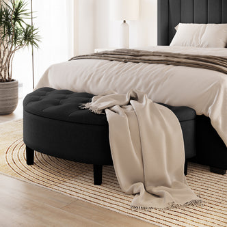 Amina Half Round Linen Fabric Ottoman in Black, tufted design, on a striped rug with a beige throw. Positioned in a modern bedroom corner, enhancing elegance and functionality.