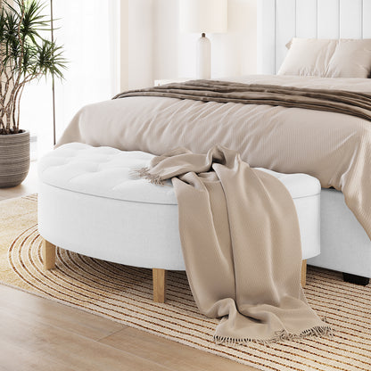 Amina Tufted Linen Fabric Ottoman in Off White, featuring a curved design with tufted upholstery and short wooden legs, positioned in a softly-styled bedroom with neutral tones and a fringed throw.