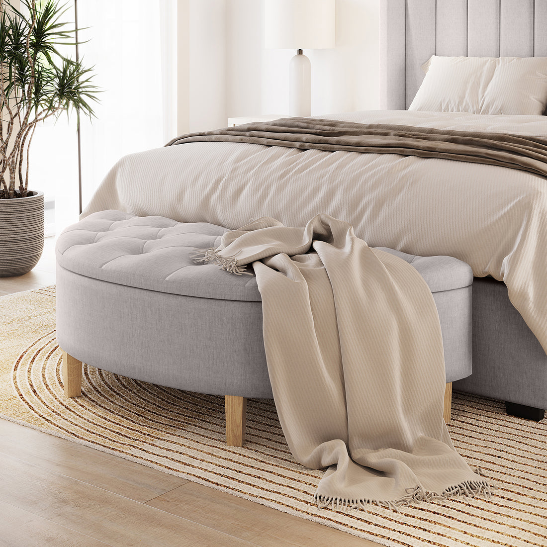 Amina Half Round Linen Fabric Ottoman in Warm Grey, featuring tufted upholstery and wooden legs, with a beige throw draped over it, positioned at the foot of a neatly made, neutral-toned bed.