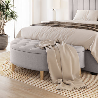 Amina Half Round Linen Fabric Ottoman in Warm Grey, featuring tufted upholstery and wooden legs, with a beige throw draped over it, positioned at the foot of a neatly made, neutral-toned bed.