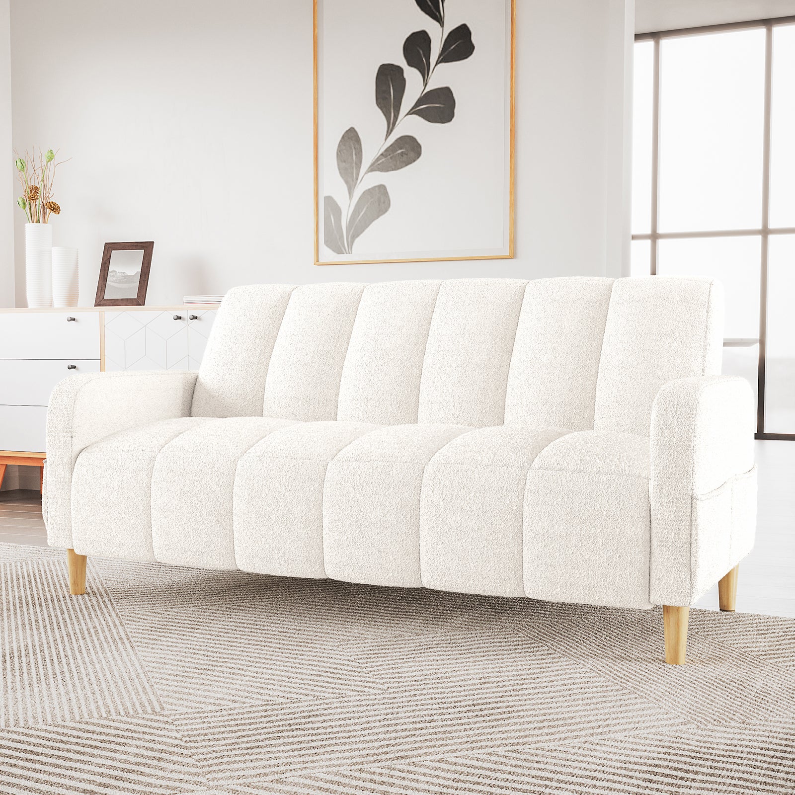 Anica 3 Seater Boucle Sofa Bed in Cream White with vertical channel tufting, light wooden legs, set in a bright room with a patterned rug, white console, and gold-framed botanical print.
