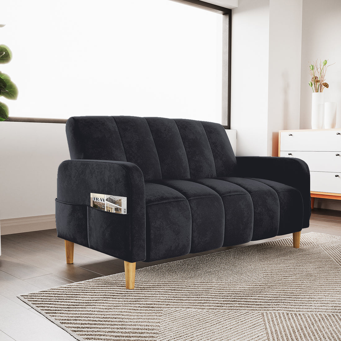 Anica 2 Seater Velvet Fabric Sofa Bed in Black with channel tufting, wooden tapered legs, and a built-in arm pocket holding a magazine, set in a cozy, sunlit modern living room.