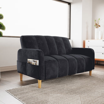 Anica 2 Seater Velvet Fabric Sofa Bed in Black with channel tufting, wooden tapered legs, and a built-in arm pocket holding a magazine, set in a cozy, sunlit modern living room.