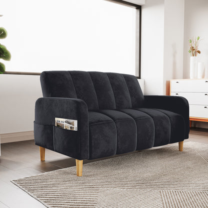 Anica 2 Seater Velvet Fabric Sofa Bed in Black with channel tufting, wooden tapered legs, and a built-in arm pocket holding a magazine, set in a cozy, sunlit modern living room.