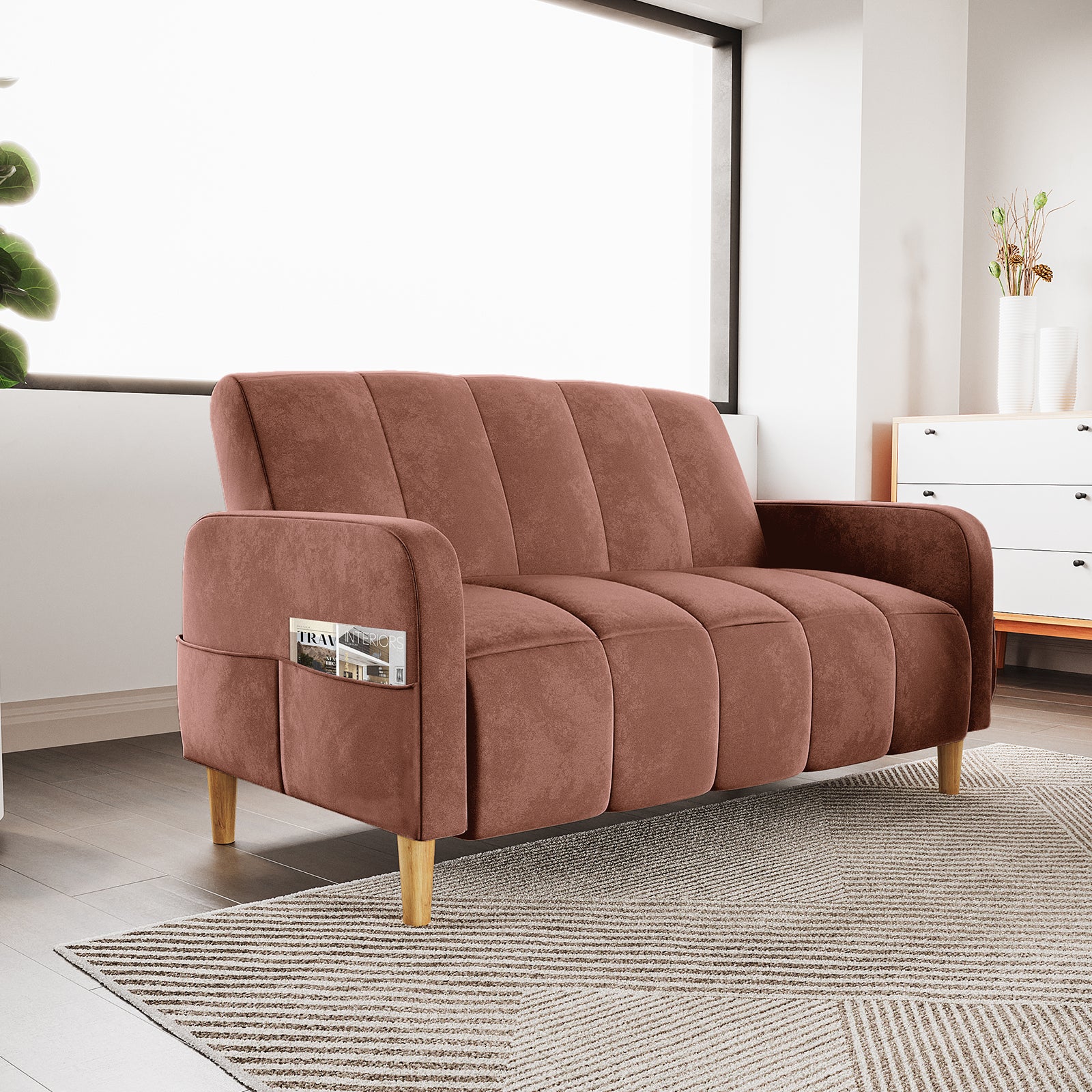 Anica 2 Seater Velvet Fabric Sofa Bed in Blush with channel-stitched cushions, slim curved arms, tapered wooden legs, and a side pocket. It sits on a patterned rug in a modern living room.