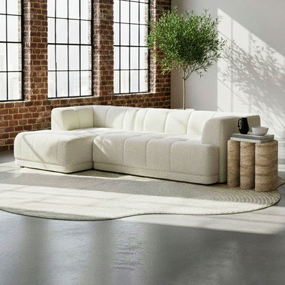 Averill 4 Seater Boucle Fabric Chaise Sofa in Light Beige, featuring a cream-white, L-shaped design with textured bouclé upholstery, set in a bright, minimalist living space with industrial-style windows and a potted tree.
