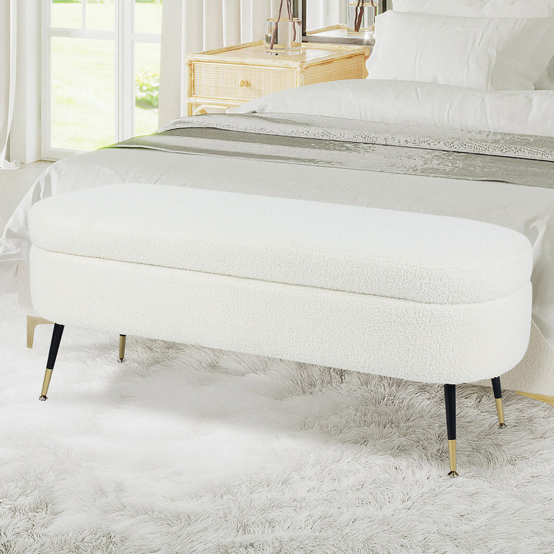 Bella 114cm Boucle Ottoman with Storage in Cream White, oval-shaped with a rounded cushion, black metal legs with brass tips, set on a fluffy area rug in a modern bedroom.