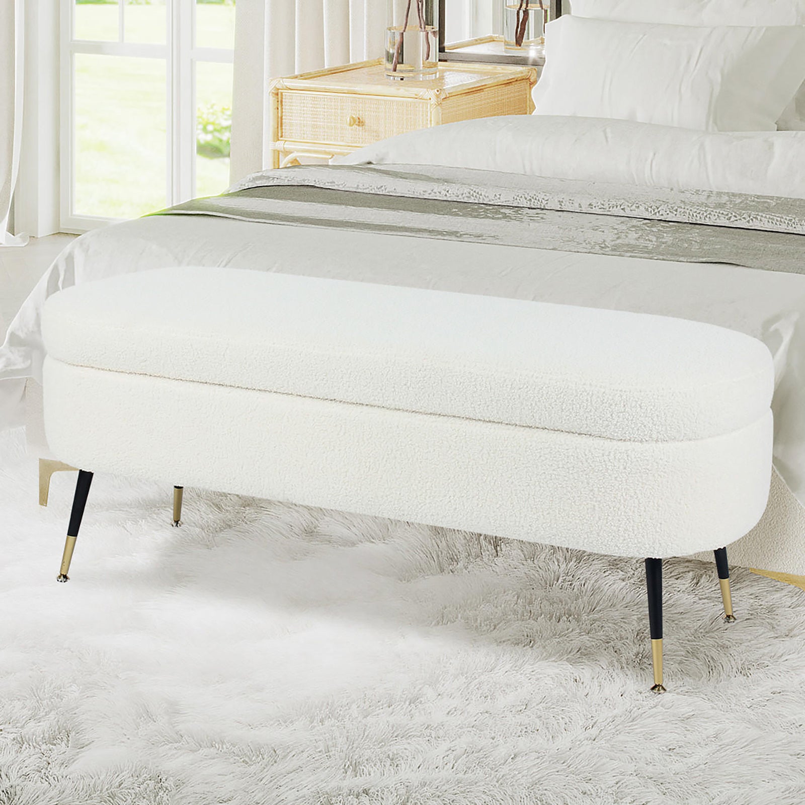 Bella 114cm Boucle Ottoman with Storage in Cream White, oval-shaped with a rounded cushion, black metal legs with brass tips, set on a fluffy area rug in a modern bedroom.
