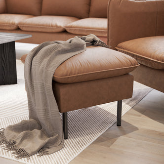 Cassian PU Leather Ottoman in warm tan leather with slender black metal legs, topped with a light-gray fringed throw, set on an off-white textured rug over light hardwood floors.