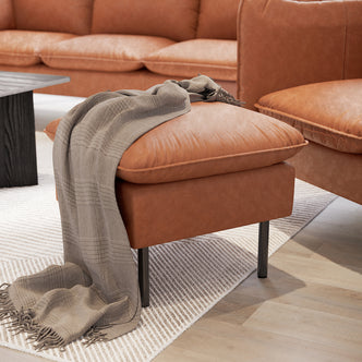Cassian PU Leather Ottoman Mocha in foreground, tan leather with a light-gray woven throw. Part of a cohesive living room set with matching chair, sofa, and black wood coffee table.